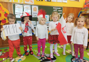 Dzieci w biało czerwonych strojach stoją jedno obok drugiego. Dziewczynka trzyma flagę, chłopiec kartkę z cyframi symbolicznej godziny 11:11.